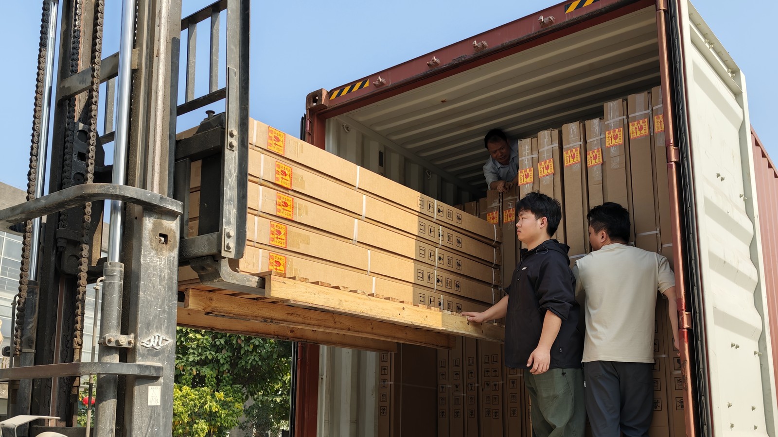 A forklift lifting a long export carton into a container while workers guide it into place